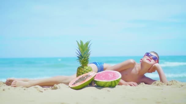 Ragazzo si trova sulla spiaggia con una composizione di frutti esotici contro il mare . — Video Stock