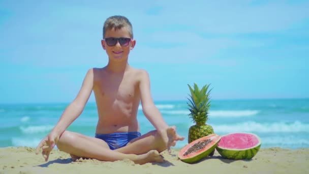 Un niño se sienta y medita en la orilla del mar cerca de frutas exóticas sobre el fondo del mar . — Vídeo de stock