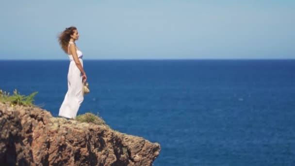 Une fille en robe blanche se tient sur une falaise rocheuse. une femme regarde au loin sur une surface de mer infinie — Video