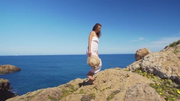 A menina sobe o penhasco descalça contra o mar azul . — Vídeo de Stock