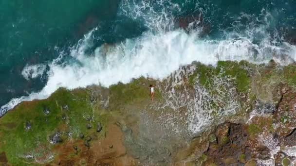 VISTA AERIAL. Las olas del mar salpicando con espuma y aerosol a la costa pedregosa. Hermosa mujer se encuentra en la playa rocosa . — Vídeos de Stock