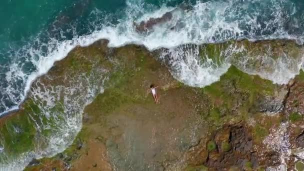 VUE AÉRIENNE. vagues de mer éclaboussant de mousse et de pulvérisation sur la côte pierreuse. Belle femme se trouve sur la plage rocheuse . — Video