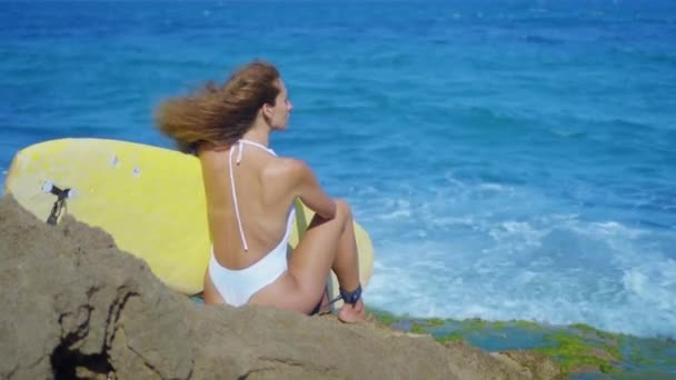 Surfer girl sits on a beautiful rocky beach with board. Powerful waves hit the rocky shore. Girl looking into distance. — Stock Video