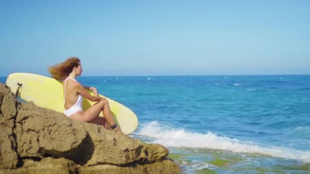 冲孩坐在一个美丽的岩石海滩上与冲浪。强大的海浪袭击了岩石海岸。女孩寻找距离. — 图库视频影像