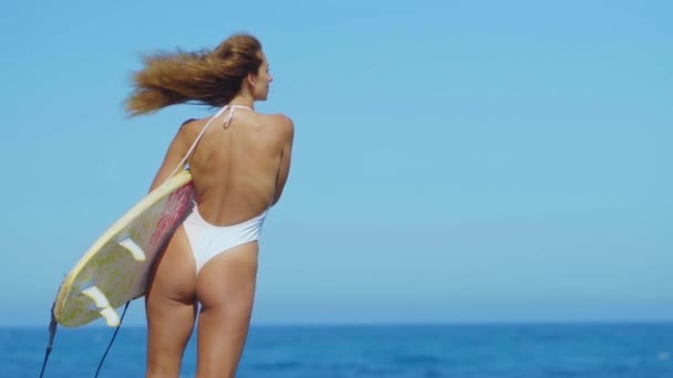 サーフボードでビーチに沿って歩いて日焼けした肌を持つセクシーな女の子。ビキニ水着で美しい若い女性。フィットネスヘルシーアクティブリビングの概念. — ストック動画