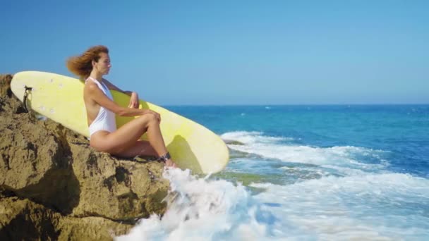 Surfermädchen sitzt an einem schönen felsigen Strand mit Brett. Mächtige Wellen treffen auf das felsige Ufer. Mädchen blickt in die Ferne. — Stockvideo