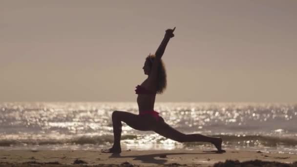 Flickan practic yoga på stranden vid solnedgången. Ung flicka gör övning på en tropisk strand. Silhuett av en kvinna yoga på en strand. — Stockvideo
