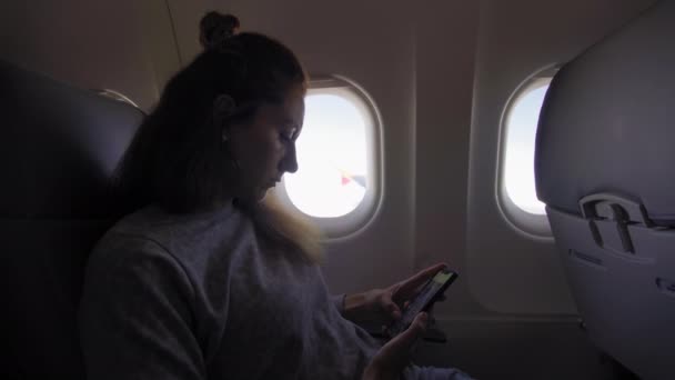 La mujer está sentada dentro de un avión, mirando en el ojo de buey y enviando mensajes de texto en su teléfono . — Vídeo de stock