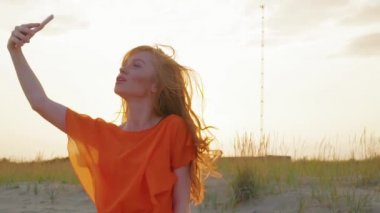 Güzel kız cep telefonu kullanarak sahilde selfie alır. Sahilde neşeli genç kadın gün batımında selfie portresi çekiyor.