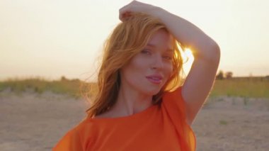 Sahilde kırmızı saçlı genç kadının portresi turuncu elbiseyle yakından karşılanıyor. Sevimli kız deniz kenarında vakit geçiriyor.