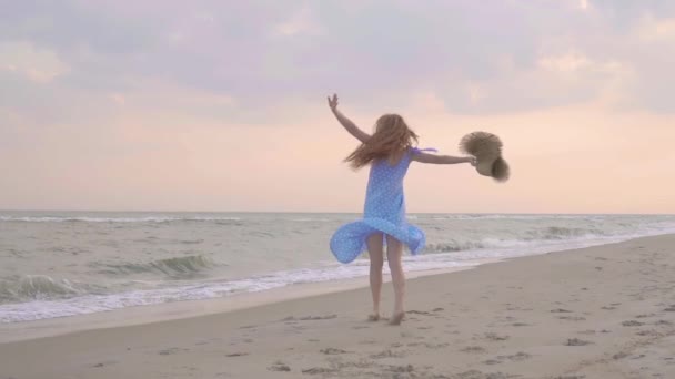 海のビーチで夕日の上で踊る気楽な女性。女の子は彼の手に麦わら帽子で回転します。夕暮れ時の女性らしさ。スローモーション. — ストック動画