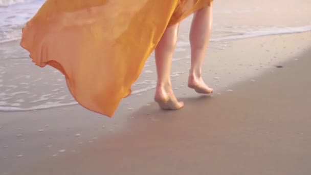 Женщины босые ноги и трепещущее платье на закате. Молодая босиком девушка в длинном оражном платье вдоль линии серфинга . — стоковое видео