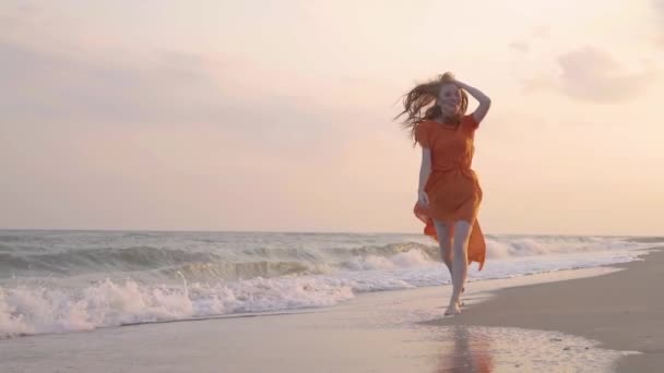 Das junge rothaarige lächelnde Mädchen läuft im orangefarbenen Kleid im Sommer am Strand am Meer. — Stockvideo