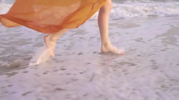 Female bare feet and fluttering dress at sunset. Young barefoot girlrunning in long orage dress along the surf line. — Stock Video