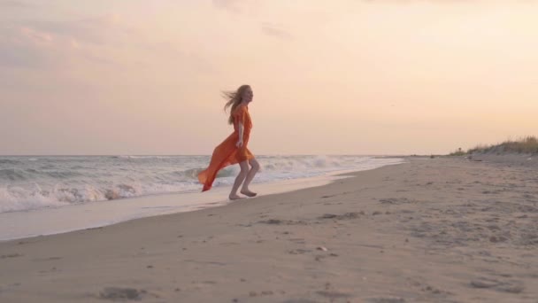 Das junge rothaarige lächelnde Mädchen läuft im orangefarbenen Kleid im Sommer am Strand am Meer. — Stockvideo