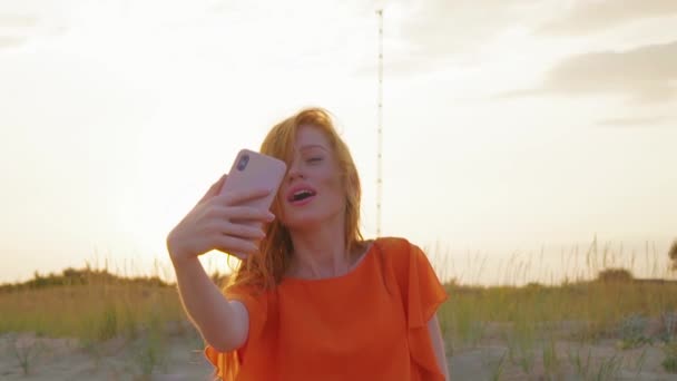 Retrato de bela jovem com cabelo ruivo tirando selfie em sua cela na praia de perto. Menina bonito gasta tempo perto do mar . — Vídeo de Stock