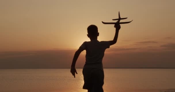 Niño feliz corre con un avión de juguete sobre un fondo al atardecer en el mar . — Vídeos de Stock