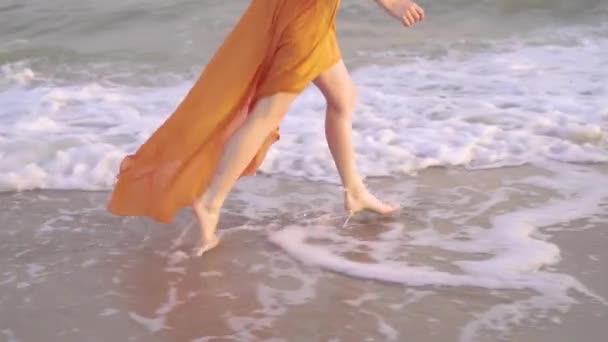 Weibliche nackte Füße und flatterndes Kleid bei Sonnenuntergang. junge barfüßige Mädels laufen im langen orangefarbenen Kleid entlang der Surflinie. — Stockvideo