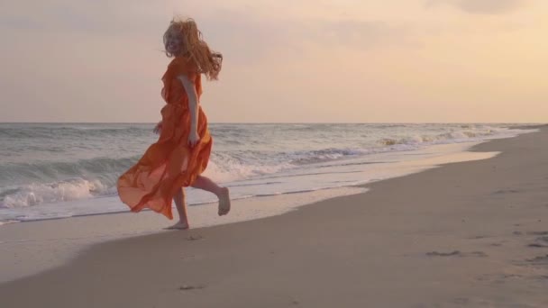 La jeune fille aux cheveux roux souriant courir dans la robe orange à la plage au bord de la mer en été . — Video