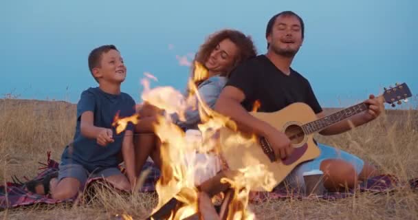 Papai toca guitarra, mãe com filho desfrutar enquanto relaxa em piquenique familiar perto de fogueira. close-up . — Vídeo de Stock