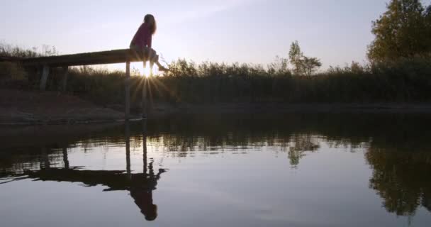 Menina bonita contempla o lago sentado no cais . — Vídeo de Stock