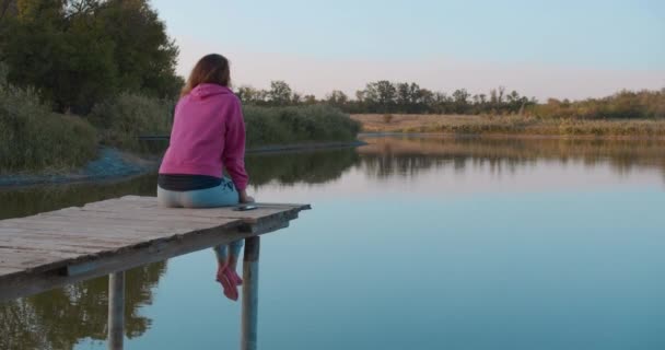 Portré egy nő ül egy fából készült mólón, nézi a tó úgy érzi, szabad. Koncepció szabadság, gyönyörű tóra néző, boldog emberek, nyaralás. — Stock videók