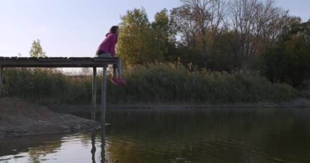 女人在湖边放松坐在一个木制码头的边缘，在夕阳上摆动腿. — 图库视频影像