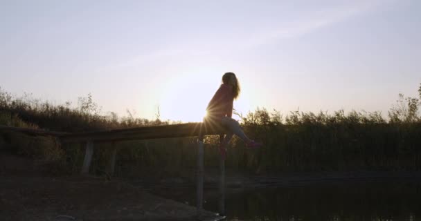 Жінка відпочиває біля озера, сидячи на краю дерев'яного дріт, розмахуючи ногами на заході сонця . — стокове відео