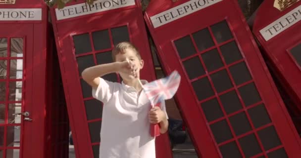 Lassú mozgás portréja Aranyos fiú integetett brit zászló áll a szabadban egyedül mosolyogva nézi kamera. A háttérben angol piros telefon fülkék. Utazás koncepció. — Stock videók