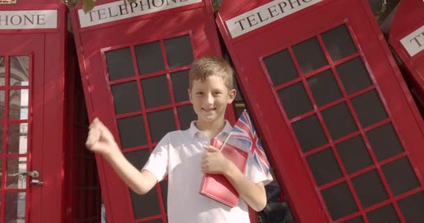 Slow Motion portret cute Boy stojący na zewnątrz sam uśmiechnięty patrząc na kamerę. Na tle angielski czerwone kabiny telefoniczne. Koncepcja podróżowania. — Wideo stockowe