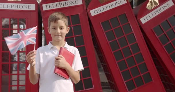 Sevimli çocuk tek başına açık havada ayakta İngiliz bayrağı sallayarak yavaş hareket portre kameraya bakarak gülümseyerek. Arka planda İngiliz kırmızı telefon kulübelerinde. Seyahat konsepti. — Stok video