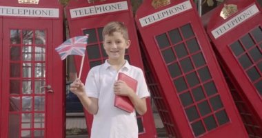 Sevimli çocuk tek başına açık havada ayakta İngiliz bayrağı sallayarak yavaş hareket portre kameraya bakarak gülümseyerek. Arka planda İngiliz kırmızı telefon kulübelerinde. Seyahat konsepti.