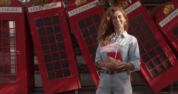 Portré lány állt a szabadban, mosolyogva nézi kamera. A háttérben angol piros telefon fülkék. Utazás koncepció. — Stock videók
