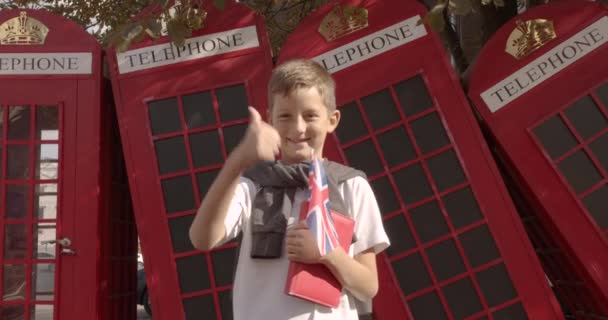 Portrait au ralenti d'un garçon mignon debout dehors, seul, souriant, regardant la caméra. Sur le fond cabines téléphoniques rouges anglais. Concept de voyage . — Video