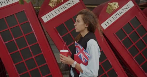 Jolie fille marchant sur un fond de téléphones britanniques rouges. Boit du café et visite touristique . — Video