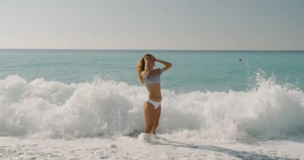 Atractiva joven mujer, traje de baño blanco se para en el mar y las olas la cubren, divirtiéndose . — Vídeos de Stock