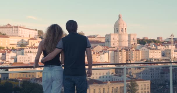 Widok z tyłu, pary obejmującej się stojąc na nasypie i patrząc na miasto Lizbona w świetle porannego słońca. — Wideo stockowe