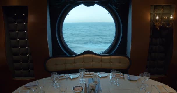 Ein servierter Tisch in einem Restaurant vor dem Hintergrund eines riesigen runden Fensters auf einem Kreuzfahrtschiff. — Stockvideo