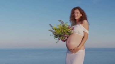 Hamile kadın denizin arka planında kır çiçekleriyle dans ediyor..