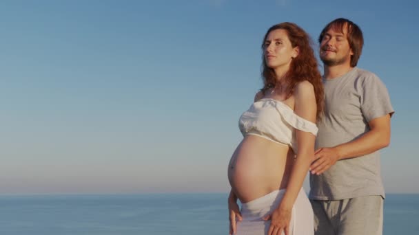 Genç hamile çift sarılıp karnını tutuyor. Anne ve baba mutlu bir şekilde gülümserken bebeğe bakarken, deniz kenarında hamilelik. — Stok video