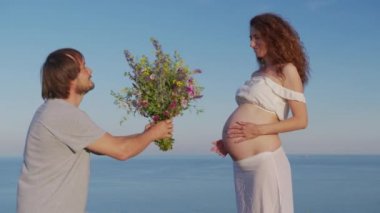 Genç hamile çift sarılıp karnını tutuyor. Anne ve baba mutlu bir şekilde gülümserken bebeğe bakarken, deniz kenarında hamilelik.
