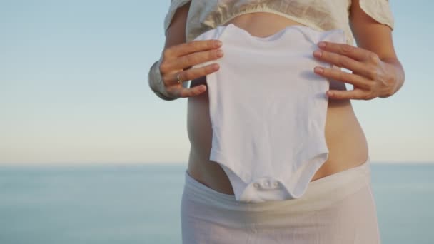 De jonge moeder die zwanger wordt en de kleren van het toekomstige jongetje op zijn buik legt. Zwangerschap prenatale zorg en vrouw zwangerschap concept. — Stockvideo