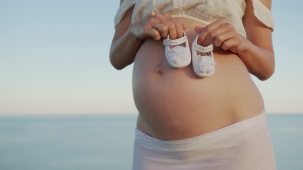 Weibliche Hände zeigen Babyschuhe auf dem Bauch. Nahaufnahme. Konzept der Neugeborenen-Schwangerschaft. — Stockvideo