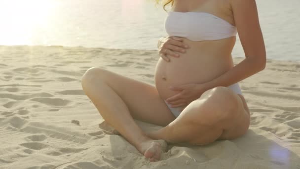 Schwangere streichelt ihren Bauch, während sie auf dem Sand am Meer sitzt. Die junge werdende Mutter hält ihr Baby im Schwangerschaftsbauch. Schwangerschaftsvorsorge und Schwangerschaftskonzept für Frauen. — Stockvideo