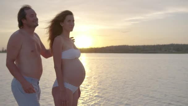 Счастливая беременная пара вместе на закате муж обнимает жену нежно держа ее живот наслаждаясь . — стоковое видео