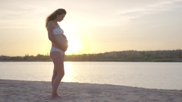 Těhotná žena drží své břicho při západu slunce na moři. — Stock video