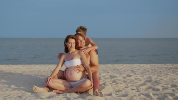 Fiatal terhes anya férjjel és kisfiával játszik és pihen a tengerparton homokkal a nyári vakáció idején. Terhesség, boldog család, szülői koncepció. — Stock videók