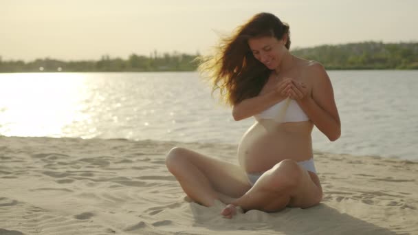 A terhes lány, aki a parton ül, homokot önt a hasára naplementekor.. — Stock videók