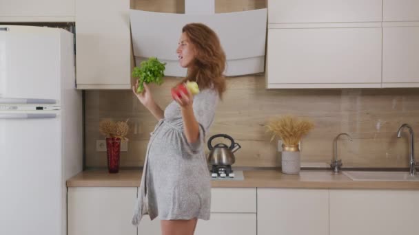 Mooie zwangere vrouw voorbereiding van voedsel en dansen met groenten op keuken. — Stockvideo