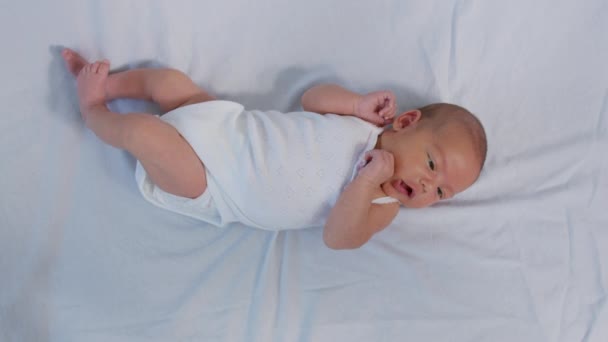 Menina recém-nascida na cama. Vista superior da linda criança em pijama branco com a boca aberta deitada na cama em movimento braços e pernas olhando para cima. Calmo bonito engraçado bebê cama vestindo pijama closeup. Infância. Hora da sesta . — Vídeo de Stock